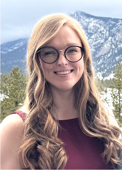 A woman with long red-blond hair and glasses is standing in front of a mountain range.