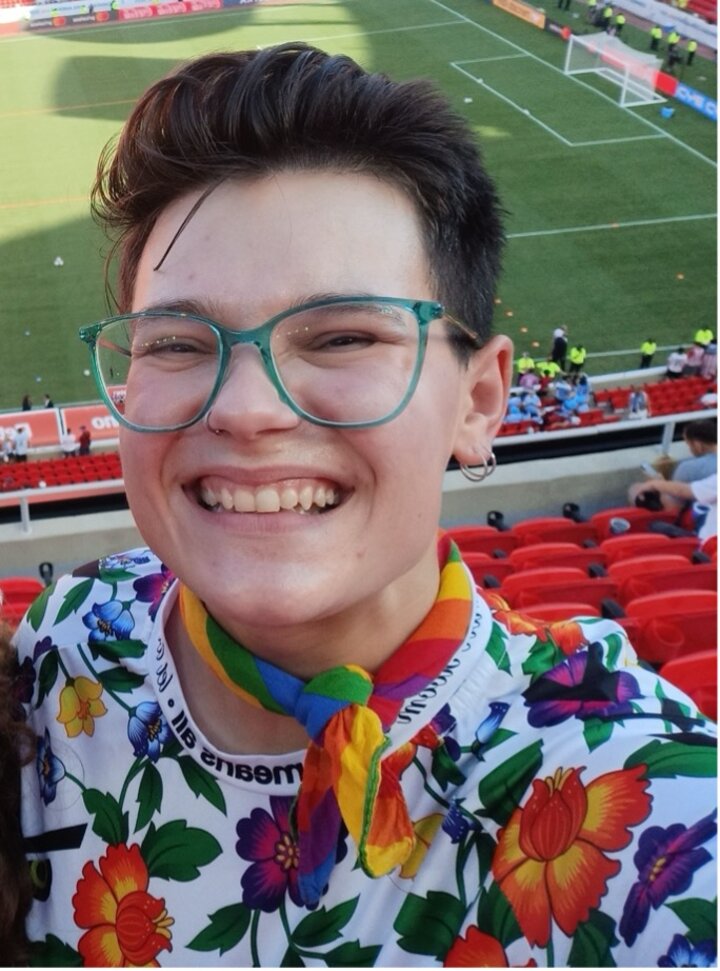 Photo of a smiling person with blue glasses, short dark hair, and a colorful shirt.